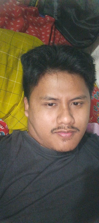 A man is pictured lying down, with his head resting on a yellow checkered cloth. He is wearing a dark gray shirt. Behind him, a red cloth with a paisley-like pattern is visible. The background also shows a black object with white text. The scene appears to be indoors, possibly in a bedroom in Kemuning, Indonesia.