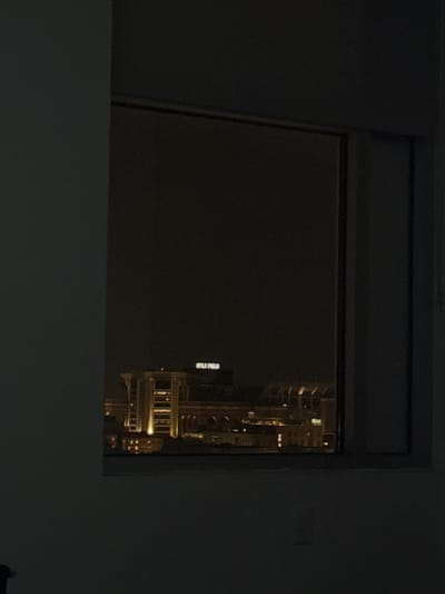 The image displays a nighttime scene viewed from inside a dimly lit interior through a rectangular window. The foreground consists of dark gray wall surfaces framing the window, with a faint light switch plate visible on the lower right wall. Beyond the dark window frame, the background reveals a brightly illuminated architectural complex, identifiable as Kyle Field stadium, located in College Station, Texas, United States of America, against a black night sky. The stadium's structures feature multiple vertical elements and arched sections, emanating a warm, yellowish-orange light from numerous internal and external sources. The name "KYLE FIELD" is clearly visible in light-colored letters on a sign atop one of the central stadium components. A small red light is discernible on an upper part of the stadium structure. No individuals, actions, or interactions are present in the scene.