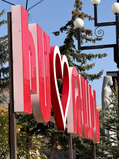 The image displays a large, three-dimensional public sign spelling "ԵՍ ❤️ ԹԱԼԻՆ" (I love Talin) in Armenian script with a central heart symbol. The sign features red frontal surfaces and white side panels, oriented diagonally from the lower left to the upper right, and is supported by dark metal posts. The background consists of a clear blue sky above and numerous evergreen trees, some exhibiting visible cones. To the right of the sign, a black metal lamppost with multiple white spherical light fixtures and decorative scrollwork is present. Further right, a light-colored statue depicting a robed figure with a head covering is visible in profile, partially obscured by foliage. The scene is an outdoor setting illuminated by bright daylight, located in Talin, Armenia. No individuals are present.