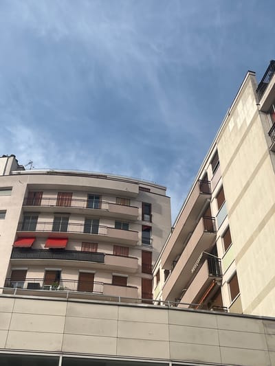 The image shows the exterior of modern apartment buildings under a cloudy blue sky. The buildings are multi-story, with visible balconies, windows, and awnings. The building on the left has a curved facade with multiple balconies featuring black metal railings. Some windows have closed brown shutters. Red awnings are visible on the lower floors of this building. The building on the right is taller and has a more angular design, with cream-colored walls featuring lighter-colored vertical stripes. Its balconies also have black railings, and some windows show brown shutters. A lower, lighter-colored building with a tiled facade is visible in the foreground, partially obscuring the lower sections of the apartment buildings. The scene appears to be in an urban environment, likely residential, in Bourg-la-Reine, France, given the context. No people are visible in the image. The overall impression is one of contemporary urban architecture under daylight.