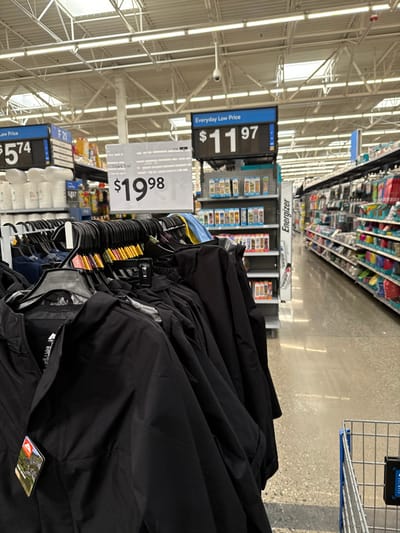 An indoor, vertically oriented image captures a retail store aisle, likely a Walmart, in Worcester, United States. The perspective is rotated 90 degrees clockwise, placing the store ceiling on the left and the floor on the right. In the immediate foreground, a rack holds numerous black jackets or outerwear garments.

Behind the clothing rack, several price signs are prominently displayed. A large blue and white sign, slightly to the left, shows "$11.97" with the text "Everyday Low Price." Directly behind the clothing rack, a white sign reads "Ozark Trail" and "$19.98." Further left and lower, another blue and white sign is partially visible displaying "$5.74" and "Everyday Low Price," next to the label "F21."

In the middle ground, store shelves stocked with various products are visible, receding into the background. A display stand features the brand "Energizer." The store has a high industrial ceiling with exposed beams, pipes, and fluorescent lighting, from which a security camera hangs. The floor appears to be polished concrete, reflecting light. No people are visible, and the scene depicts a typical shopping environment during daylight hours, illuminated by artificial lighting.