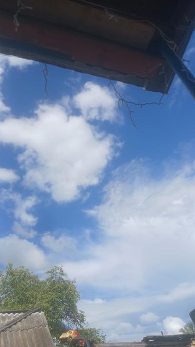 The image depicts a cloudy blue sky with a view looking upwards, suggesting it was taken from a low vantage point. In the foreground, part of a wooden structure with some hanging string lights is visible, possibly an overhang or a roof. Below this, to the left, is a gray, corrugated roof. To the right of the corrugated roof, a vibrant green tree with dense foliage is present, with a small yellow and orange toy truck visible amongst some debris, possibly on a tire, in front of the tree. The overall scene suggests an outdoor environment, possibly a backyard or a less developed area. The image does not provide enough information to determine the specific location within Slak, Russia, or to identify any individuals or specific actions taking place.