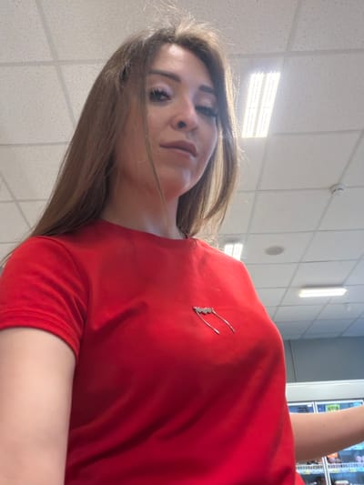 The image shows a young woman with long brown hair, wearing a red t-shirt. She has makeup on, including eyeshadow and mascara. There is a silver-colored pin on her t-shirt, which appears to be shaped like a lightning bolt or a stylized wing. The background suggests an indoor commercial setting, possibly a store, with a tiled ceiling and fluorescent lighting visible. There are also glimpses of refrigerated display cases in the background, further suggesting a retail environment. The lighting in the image is bright, and the perspective is from a low angle, looking up at the woman. The location is identified as Nurkeevo, Russia.