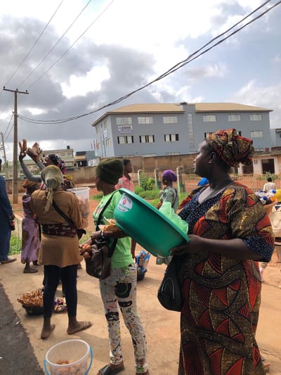 The image depicts a street scene in AlakIa, Nigeria. A group of people are gathered in what appears to be a marketplace or informal trading area. A woman wearing a patterned dress and headwrap is holding a green plastic basin. Another woman in a green shirt and patterned leggings stands nearby. Several other individuals are present, some holding food items like bread and eggs. The environment includes a building with "GOSHEN HIGH SCHOOL" signage, overhead power lines, and a paved surface. The sky is overcast.