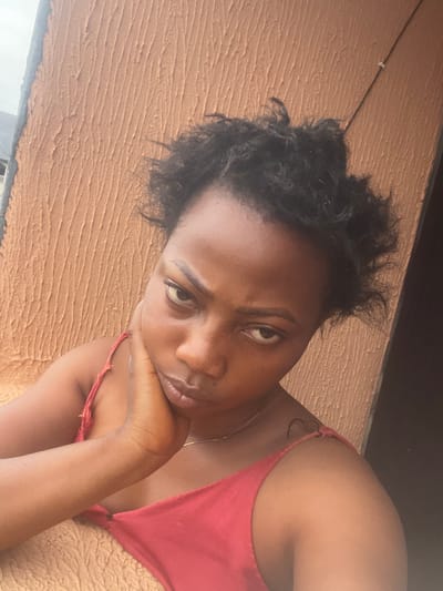 A young Black woman with curly black hair and brown skin is captured in a close-up, head-and-shoulders selfie. She is wearing a red tank top and resting her chin on her hand, with a contemplative or slightly upset expression. The background is a textured, ochre-colored wall, characteristic of the outdoor walls found in Iyiowa Odekpa, Nigeria. The lighting suggests it is daytime.