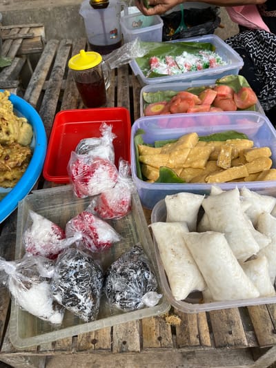 An outdoor food stall in Tambakreja, Indonesia, displays a variety of snacks on a wooden surface. In the foreground, to the left, a blue bowl contains battered and fried food items. Next to it, a red tray holds several plastic-wrapped snacks, some appearing red and white, and others dark brown with white flecks. Further to the right, two clear plastic containers are filled with different types of food. One contains rectangular, golden-brown pieces covered with banana leaves, while the other is filled with white, rice-like packages. In the background, a glass mug with a yellow lid holds a dark beverage, and a jug with a tap is also visible. The stall is set up on a rustic wooden surface, likely a table or a series of planks, with some foliage and other items in the periphery. The scene appears to be a casual market or roadside setup, typical of street food vendors in Indonesia.