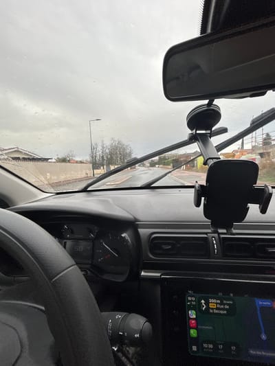 The image presents a forward-facing view from inside a vehicle located in Saint-Cyprien, France, looking out through a windshield covered with raindrops. The foreground is dominated by the vehicle's interior: the left portion of a steering wheel is visible on the bottom left, partially obscuring the instrument cluster which displays "50 4" and a gauge. A black smartphone holder is mounted via suction cup to the windshield and extends downward, positioned over the central dashboard air vents. An infotainment screen below the vents displays a navigation application showing "10:13," a "5G" network indicator, a direction instruction for "300m" to "Rue de la Becque," and an estimated arrival time of "10:30." A rearview mirror is visible at the top center.

Outside the vehicle, the road ahead is asphalt and appears wet. Windshield wiper blades are visible in an upward-swept position across the lower windshield. The environment is a suburban or residential street lined by low fences, walls, and houses with tiled roofs. Deciduous trees, devoid of leaves, stand further down the road, accompanied by a streetlamp. On the right side of the road in the mid-distance, building materials and scaffolding are visible, suggesting a construction site. The sky is uniformly overcast and grey, indicating rainy or recently rainy weather conditions. No individuals are visible in the scene.
