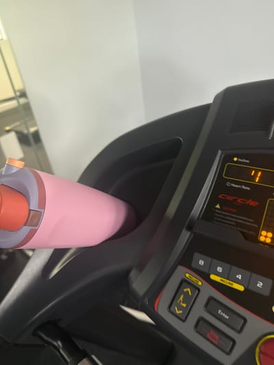 The image depicts a close-up, angled view of a black 'Circle' brand treadmill console and its integrated cup holder, situated within an indoor fitness facility in Hallandale Beach, United States. The precise address or name of the facility is not discernible from the visual information. A pink water bottle, featuring a grey mid-section, reddish-orange mouthpiece, and a small bronze detail, is horizontally positioned in the cup holder on the left. The treadmill's digital display indicates an 'Incline' of '1' and shows a 'Heart Rate' field with no value, alongside four illuminated orange squares. Visible controls include numeric buttons, 'Incline' adjustments, 'Enter,' and 'Stop/Reset.' The background features a plain, light-colored wall, with a blurred reflection of an interior space, possibly a mirror or glass partition, visible in the upper left. No individuals are present in the frame.