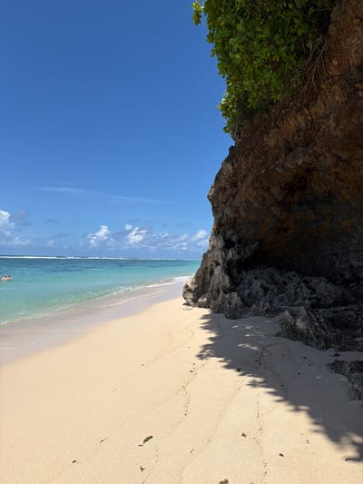 The image captures a beach scene in Benoa, Indonesia. The view is oriented with the sea and sky on the left and the sandy beach on the right. The sky is blue with scattered white clouds. The ocean transitions from turquoise near the shore to a deeper blue further out. A small wave breaks along the shoreline. In the distance, two figures are visible in the water.

A rocky cliff overhangs part of the beach at the top of the frame. Green vegetation grows on the left side of the cliff. The sand is light-colored with some debris scattered across it. Shadows from the vegetation on the cliff fall onto the sand.