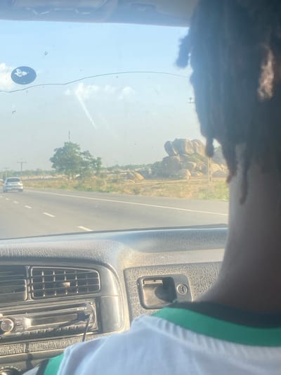 The image shows a view from inside a vehicle, likely a car, looking out through the windshield. The vehicle is moving along a paved road.

**Location:** Unguwar Fatika, Nigeria is inferred from the environment visible outside the vehicle. The landscape includes rocky hills or formations, sparse vegetation, and power lines.

**People:** A portion of a person's head is visible in the upper left of the frame. They have dark hair, possibly in dreadlocks. Part of another person's shoulder, covered in a white and green garment, is visible on the right side of the frame.

**Objects:** The interior of the car is visible, including the dashboard, air vents, a Sony radio/cassette player, and a door lock/handle. A butterfly shaped sticker is visible on the front windshield. A vehicle is traveling on the road ahead.

**Environment:** The scene is outdoors on a sunny day. The road appears to be well-maintained.