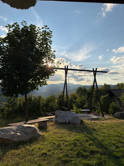 The image depicts an outdoor scene, likely a park or backyard, in the city of Trebishte, Bulgaria. In the foreground, a large, rough-textured boulder sits on a grassy area bathed in the warm glow of late afternoon sunlight. To the left, a wooden bench with concrete supports is positioned on a patch of dirt and grass, with another large boulder to its right. Dominating the midground is a wooden swing set with a wooden bench suspended by metal chains. Behind the swing set, a dense line of trees stretches across the landscape. Further in the background, rolling hills, covered in vegetation, recede into the distance, creating a layered mountainous panorama. A small, dark-colored building with a pitched roof is partially visible on the right side of the frame, nestled amongst the trees. The sky above is a vibrant blue, adorned with scattered white clouds, some of which are illuminated by the setting sun, casting light rays through the atmosphere. A large tree with dark green foliage is situated on the left, its branches partially obscuring the sun, creating a lens flare effect. The overall impression is one of a serene, natural setting at dusk.