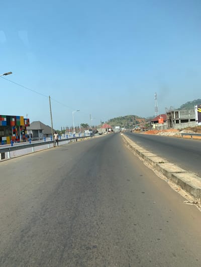 The image, rotated 90 degrees counter-clockwise from its original orientation, depicts a long, straight asphalt road extending into the distance under a clear blue sky. The scene suggests a developing area, possibly a highway or main thoroughfare in Tokeh, Sierra Leone, given the provided context.

The road itself is a two-lane expanse of asphalt, showing some wear and tear but generally appearing functional. On the right side of the road, a concrete barrier or curb separates the pavement from an unpaved shoulder. Beyond this, several buildings are under construction, characterized by exposed concrete frames and rebar. One building features partially completed walls and a large, dark sign with visible yellow letters "ABU", possibly part of "ABUJA" or a similar name, attached to its upper level. Further down, a tall communications tower or mast is visible, accompanied by a smaller red sign with unreadable white text. Hills rise in the background on this side, indicating a varied topography.

On the left side of the road, a metal guardrail runs alongside the asphalt. In the foreground, a distinctive, low barrier or wall composed of stacked, multi-colored rectangular blocks (blue, green, yellow, orange, red, black) creates a vibrant visual element. A single person is visible standing near this colorful barrier, facing away from the camera, wearing light-colored clothing. Numerous streetlights with modern fixtures line this side of the road, extending into the distance at regular intervals. Beyond these, more construction sites with unfinished buildings are present, mirroring the development seen on the opposite side.

The time of day appears to be daytime, with bright sunlight and a cloudless, clear blue sky, suggesting fair weather. The overall impression is one of ongoing infrastructure and urban development in a natural, hilly landscape.