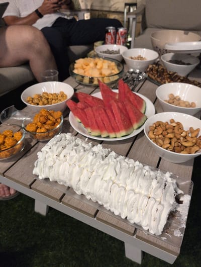 This is a medium shot, eye-level, horizontal photograph taken outdoors at dusk. The scene depicts a table laden with snacks and a dessert. In the foreground, a long, rectangular roll cake covered in white frosting is positioned horizontally. The cake appears to be a chocolate roulade, with visible hints of dark chocolate peeking through the frosting. To the left of the cake, three small glass bowls are visible, filled with what appear to be assorted fried snacks. Further to the left, a person's legs and feet are visible, resting on a couch, with one foot bare and another wearing a sandal.

The middle ground of the image showcases various bowls of snacks arranged on a wooden table. A white plate holds several triangular slices of watermelon. Next to the watermelon, a bowl contains what looks like dried fruit, possibly apricots or mangoes. Other bowls contain an assortment of nuts, including cashews and possibly peanuts. Two cans of Coca-Cola are visible in the background. The table is set on an artificial turf surface, suggesting an outdoor gathering. The overall setting implies a casual get-together or party. There is no indication of the specific location within the city of Lod, Israel, in the image itself.