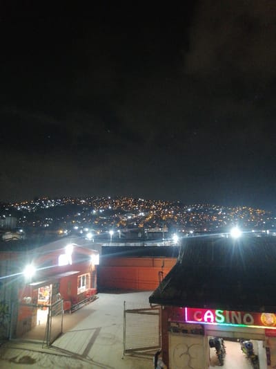 A nighttime cityscape is seen, with numerous lights illuminating a hillside. In the foreground are two buildings. The building on the right has a "CASINO" neon sign. To the left is a smaller, brightly lit shop. A fence separates the buildings. The sky is dark with some cloud cover, and a few faint stars are visible. The scene is captured from an elevated perspective.