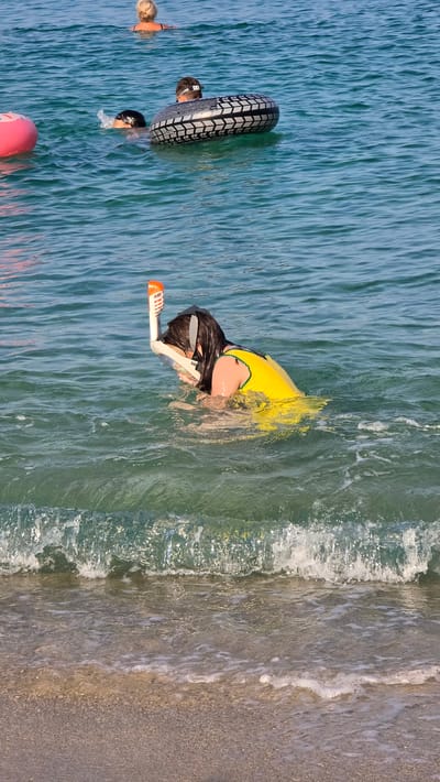 A person wearing a yellow life vest and snorkel gear is swimming in the ocean near a beach. The person's face is partially submerged in the water. In the background, another person is swimming with a tire-shaped float. In the distance, a third person is visible further out in the water. The water is clear and blue, with small waves rolling towards the sandy shore. This scene is located in Toroni, a municipal unit in Chalkidiki, Greece, known for its beautiful beaches and clear waters.