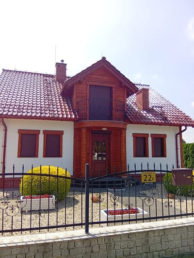 The image shows the exterior of a two-story house located at ul. Lipowa 22F in Brzezia Łąka, Poland. The house has a white facade with red-tiled roofing and wooden accents. It features a prominent wooden entrance with a balcony above it, two windows on the lower level with dark shutters, and a brick chimney. A black wrought-iron fence encloses the property, with a stone wall at the base of the fence. Inside the fence, there are gravel pathways, a yellow bush, and white planters with red flowers. A brown mailbox is attached to the fence. The sky is clear and blue. There are no people visible in the image.