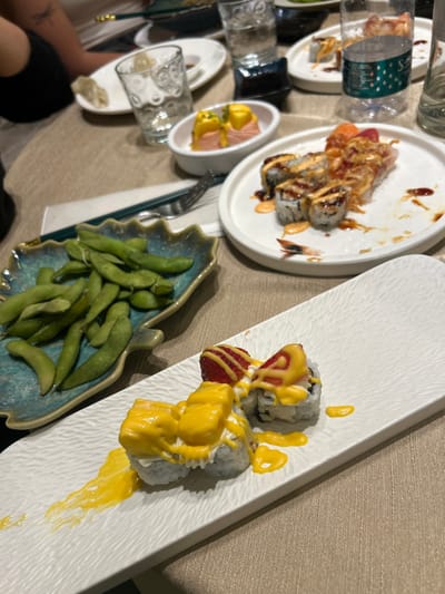 The image depicts an indoor dining scene at a restaurant in Borgo San Dalmazzo, Italy, showcasing various dishes and tableware on a light-colored table surface.
In the foreground, a rectangular white plate with a textured surface holds four sushi rolls. Each roll is topped with a vibrant yellow sauce, and two of the rolls are further garnished with sliced strawberries. To the left, a turquoise, leaf-shaped ceramic dish contains a pile of green edamame pods.
In the mid-ground, a round white plate displays several partially consumed sushi rolls with various toppings, including what appears to be crispy fried onions, and drizzles of dark and orange sauces. A small, cooked prawn tail is visible on this plate. Behind it, a small oval white dish contains two portions of food topped with a similar yellow sauce.
Further into the background, a person's upper arm and shoulder, wearing a dark garment, are visible on the left, with a faint tattoo resembling a dragonfly on the bicep area. Multiple clear drinking glasses, some containing water, are distributed across the table. One glass has a distinct patterned surface. A plastic water bottle with a blue-green label is also present. A set of dark green chopsticks and a silver fork rest on a white napkin between the edamame dish and the round sushi plate. Another white plate in the background holds a single dumpling. The overall arrangement indicates a dining setting with multiple dishes for consumption.