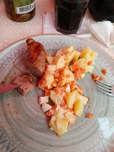 The image shows a plate with a partially eaten meal of potato salad with diced carrots, hard boiled eggs, and a sausage. There are remnants of the meal on the plate, suggesting it has been partly consumed. The background shows a partially full glass of cola, an olive oil bottle, and a white napkin. This appears to be a casual dining experience, with the meal likely consumed at home or in a casual setting. The location of this scene is El Talar, Argentina, based on the provided coordinates.