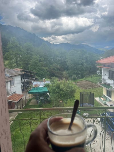 A first-person perspective shot captures a glass of coffee with a spoon in it, held by a hand in the foreground. The background reveals a view from a balcony overlooking a rural village nestled within a valley. Houses with tiled roofs are scattered among lush green trees, with agricultural fields visible in the mid-ground. Majestic mountains form the backdrop, with a cloudy sky overhead. The location is identified as Elhovets, Bulgaria.