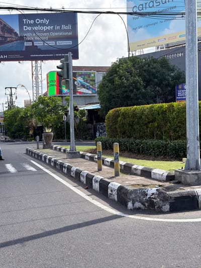 An eye-level, full shot shows a street intersection in Denpasar Barat, Indonesia, under an overcast sky. The foreground features a paved road with a white pedestrian crossing. The middle ground showcases a concrete median with black and white striped borders, and a traffic light with green and red signals. Beyond the median, a well-maintained green hedge lines the sidewalk, interspersed with trees. In the background, billboards advertise real estate and sunscreen. The scene captures a typical urban environment with its blend of infrastructure, advertising, and lush vegetation.