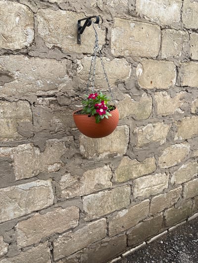 A medium shot shows a hanging planter with vibrant pink flowers against a rough, textured stone wall. The planter is suspended by a metal chain from a black metal bracket affixed to the wall. The wall is constructed from large, irregularly shaped stones with visible mortar. The ground beneath is covered in dark gravel. The lighting suggests an outdoor setting, likely during the daytime. The scene appears to be in the city of Kinel'-Cherkasy, Russia.