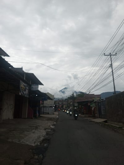 The image displays a street view in Sindanglaya, Indonesia, under an overcast sky. A narrow, paved road extends into the background, bordered by single-story buildings on both sides. Structures on the left exhibit corrugated roofs and multiple storefronts, some secured by closed metal roll-down shutters. On the right, a utility pole supports numerous overhead electrical wires. In the distant background, a mountain peak is visible, partially obscured by clouds. A procession of at least five motorcycles, with illuminated headlights, proceeds away from the viewer along the road. The lead motorcycle rider is observed wearing a helmet. The immediate foreground of the road's edges shows uneven pavement and scattered debris.