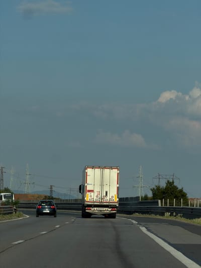 A white semi-trailer truck and a dark-colored car are traveling on a highway in Yana, Bulgaria. The truck is in the foreground, and the car is behind it. The highway has a white line marking the lanes. In the background, there are power lines and trees. The sky is cloudy. The sun appears to be high in the sky, suggesting it is daytime. The image is taken from a slightly elevated perspective, looking down the highway. There is no clear indication of emotions or a specific event taking place beyond normal highway travel.

Visible text includes "80" and "90" on the back of the truck, likely indicating speed limits or cargo information. The license plate on the car is partially visible, showing "BP 8351 CT". The truck appears to be a semi-trailer refrigerated truck, indicated by the white insulated box body. The car is a hatchback. The power lines are a prominent feature of the landscape, typical of rural or semi-rural areas with high voltage transmission. The overall scene is one of a typical day on a Bulgarian highway.