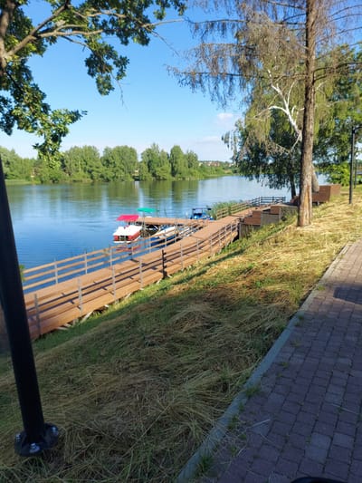 This media file depicts a tranquil waterside scene. The main subjects are a wooden boardwalk along a calm body of water, lush green trees lining the opposite bank, and a clear blue sky overhead. Several small boats are docked at the boardwalk, some adorned with colorful cloths, suggesting a recreational area. The grass along the boardwalk is dry and yellowish, indicating it might be late summer or early autumn. A paved pathway runs alongside the grassy area, bordered by dark grey bricks. The overall impression is one of peacefulness and natural beauty. The time of day appears to be late afternoon, given the angle of the sunlight creating shadows. There is no visible text in the image.