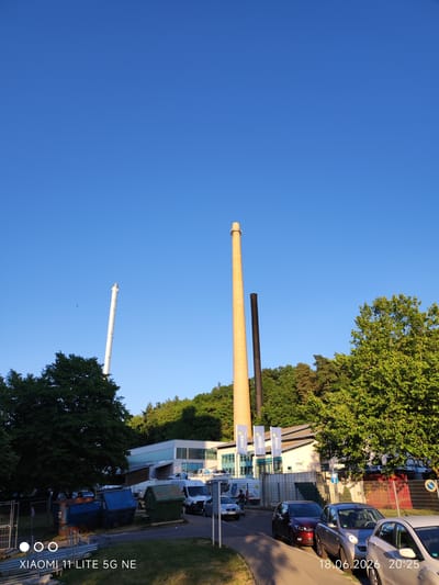 This image shows an industrial area in Böblingen, Germany, likely a former or active power plant, on a clear day. Dominating the scene are two tall chimneys: a taller, older brick chimney to the right and a thinner, white, modern-looking chimney to the left, partially obscured by trees. In the foreground, several large industrial dumpsters are visible. A row of cars is parked along a street, and a white van is also present. Behind the dumpsters and cars, a low-rise industrial building with large windows and a light-colored facade stands. Lush green trees and foliage surround the area, with a dense forest visible in the background on a sloping hill. The sky is a vibrant, clear blue. The timestamp in the bottom right corner indicates the photo was taken on June 18, 2026, at 20:25. The text "XIAOMI 11 LITE 5G NE" in the bottom left corner is a device watermark.