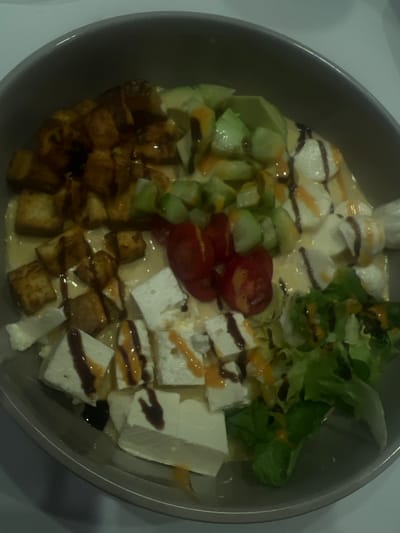 A top-down view reveals a bowl containing a salad with distinct sections. Cubed, dark-colored, possibly fried tofu occupies the lower left. Adjacent are sliced cucumbers and halved cherry tomatoes. Mozzarella cheese is located in the top-center. Crisp lettuce leaves are placed at the upper right. Cubes of feta cheese fill the lower right section. Drizzles of what appears to be balsamic glaze and another lighter colored sauce are scattered over the ingredients. A light-colored dressing or sauce is seen coating the bowl's base.
