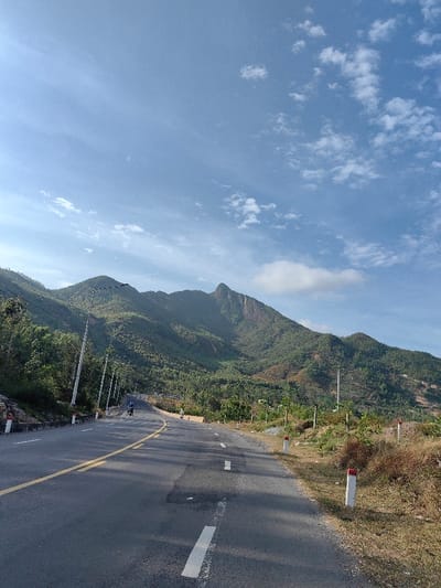 The image depicts a two-lane asphalt road curving gently uphill to the left, set within a mountainous landscape in Tuy Phong 4, Vietnam. The road features a double solid yellow centerline and white dashed shoulder lines, with areas of darker asphalt indicating repairs. White and red reflective road markers (bollards) line both sides of the road.
On the left side of the road, a series of utility poles with overhead wires ascend alongside a concrete barrier. Beyond these, the mountain slopes are covered with dense green foliage. In the mid-distance on the road, two dark-colored motorcycles, each with a single rider, travel away from the viewer. The lead rider wears a light-colored helmet.
The right side of the road transitions to a dry, earthy shoulder with sparse, dried vegetation, leading to more green trees and shrubs covering the mountain slopes. In the background, prominent green mountains dominate the scene. The highest peak features a distinct, rugged exposed rock face. The sky above is clear blue with scattered white cumulus and cirrus clouds. Sunlight illuminates the scene, indicating daytime conditions.