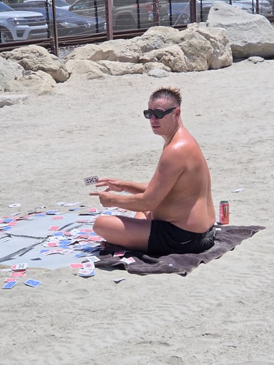 A shirtless man wearing black swim trunks and sunglasses is sitting on a towel on a sandy beach in San Clemente, California. He is surrounded by a large number of playing cards scattered on the sand. The man is holding one card in his left hand and appears to be in the process of placing it down or throwing it. A can of Monster energy drink is visible next to the towel. In the background, there are large rocks and a fence with vehicles visible behind it. The overall impression is of a sunny day at the beach with recreational activity.