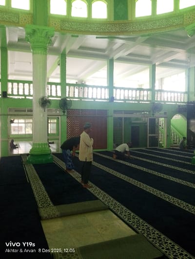 Inside a mosque, dark blue carpets with ornate borders stretch across the floor. Several men are present, some standing, others kneeling in prayer, all facing a direction not visible. One man stands with his arms crossed. A large, green column supports a balcony with a balustrade. Islamic calligraphy adorns the upper walls. Fans are mounted on the walls. Sunlight filters through windows, illuminating the interior. A partially rolled carpet lies in the foreground. A watermark is at the bottom left.