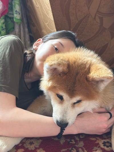 A woman lies on what appears to be a bed or sofa, with a tan and white Akita dog resting on her chest and arm. The woman has dark hair and is wearing a green T-shirt. The dog's face is close to her face. The location is Заболотово, Russia, but this is not verifiable from the image. The surrounding environment includes a patterned blanket or cushion and what looks like a piece of furniture with a brown pattern. The woman's hand is visible, wearing a black ring.