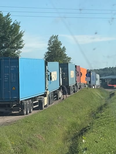 The image displays a line of semi-trailer trucks, their containers stacked high, suggesting a traffic jam. The trucks are parked along a grassy roadside, with a dirt path or shoulder visible between the road and the grass. In the background, a dense line of trees forms a natural boundary. The sky is a clear blue, with a few wispy clouds, indicating fair weather. The overall scene conveys a sense of standstill, with the trucks lined up as far as the eye can see.

The time of day appears to be daytime, with bright sunlight illuminating the scene. The location is identifiable as Zarech'e, Russia. The photo seems to be taken from a vehicle, possibly another car or truck, as indicated by the angle and slight blurriness, giving a sense of being part of the traffic.

There is some visible text on the containers. On the blue container in the foreground, the text "FITU 568730 0 45G1" is clearly legible. There is also a yellow triangle with a black symbol inside on this container. Other containers have various markings, but they are less distinct. The scene suggests a common occurrence of traffic congestion, possibly due to road work or an accident, although no specific cause is evident. The focus is on the logistical aspect of transportation and the resulting traffic flow.