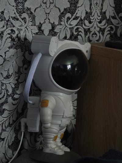 The image shows a white astronaut-shaped lamp with a black helmet, standing on a dark surface. The lamp is positioned to the left of a wooden shelf, and behind it is a black and white patterned wallpaper. A white cable is connected to the back of the lamp. The scene is dimly lit. There is no discernible urban environment or specific location within Зажэвіцкі сельскі Савет, Belarus, identifiable in the image. The description is limited to the visual content of the photograph.
