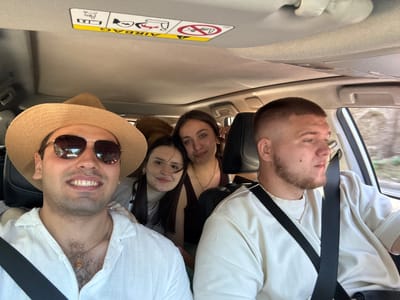 Four people are inside a car, appearing to be on a road trip. In the foreground, a man wearing a straw hat and sunglasses smiles at the camera, dressed in a white linen shirt. Beside him, another man with a short haircut and beard is looking to the side, wearing a light-colored sweatshirt. Behind them, two young women are seated, with one in the middle looking towards the camera and the other in the back with her head slightly tilted. The interior of the car shows the ceiling with an airbag warning sticker. The background is blurred, suggesting movement and the exterior environment is not clearly discernible, but the location is identified as Skala Rachoniou, Greece.
