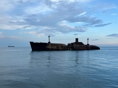 A forensic examination of the image reveals a wide shot of the Black Sea off the coast of Costinești, Romania, featuring the prominent shipwreck of the cargo ship Evangelia. The vessel, painted dark blue/black and extensively rusted, is visibly broken into two primary sections, with a significant gap near its midship. The forward section displays "EVANGELIA" painted in yellow/orange letters on its hull, beneath which "VARA CEA MAI FIEBNE R GASTITATE" is also visible. The stern section shows graffiti that reads "Summer Camp, U.S.R." Birds, likely gulls or cormorants, are perched on various structures of the derelict ship, including the mast remnants and the bridge superstructure. Several birds are also seen flying near the wreck. The sea is calm, with subtle ripples on its dark blue-green surface. In the distant background on the left, another maritime vessel, possibly a barge with a crane, is faintly visible on the horizon. The sky is partially overcast with a mix of light blue and scattered greyish-white clouds. No human figures are present in the immediate vicinity of the shipwreck.