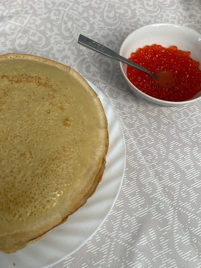 This image depicts a domestic table setting in the city of Пугачёво, Russia. A stack of approximately five golden-yellow blinis (Russian pancakes) is presented on a white ceramic plate with a subtly fluted edge, occupying the lower-left and central portions of the frame. To the upper right, a small white ceramic bowl with a pale blue decorative band contains a significant quantity of red caviar. A silver-colored spoon, bearing the inscription "G. I. L. L. A. I. T." on its handle, is partially submerged in the caviar. The entire arrangement rests on a light-colored tablecloth patterned with a repeating design of small, interconnected dots forming abstract geometric shapes. No individuals are present in the scene.