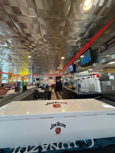 Here is a detailed description of the image:
**Location:** The image appears to be taken inside a diner or restaurant. The environment suggests that it is located in a busy commercial area, likely within a city. The OCR text indicates that the restaurant might be in West Ocean City, United States of America, as it mentions "BEACH21!" and "Francis Scott Key Guest" which is relevant to this location.
**Environment:** The restaurant has a long counter with a polished metal wall behind it, decorated with a pattern of squares, each with a smaller diamond-shaped pattern inside. Above the counter, there's a row of glasses and bottles, indicating a bar area. There are TV screens displaying sports or news. The ceiling is illuminated with bright lights, and there's a red light strip running along the upper wall.
**Objects and Subjects:** In the foreground, there is a box of Jim Beam whisky. The restaurant has a Pepsi sign. There are straws and some food items placed along the counter. There appears to be a customer or worker at the counter.