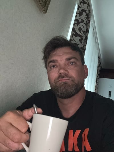 The image is a close-up, rotated selfie of a man holding a white mug. He appears to be indoors, in what looks like a bedroom or living area. The background shows blinds with a patterned curtain, and part of a white appliance with the text "INDESIT" visible. The man has brown hair, a beard, and is looking directly at the camera with a neutral expression. He is wearing a black t-shirt with large orange letters that appear to spell "KKAA". The lighting suggests it could be daytime, possibly late afternoon or evening. There are no clear indicators of the weather or specific time of day beyond that. The overall impression is casual and personal.