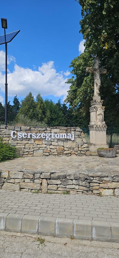 The image shows a sign with white lettering that reads "Cserszegtomaj" on a stone wall. To the right of the sign is a stone cross with a depiction of Jesus Christ and another figure at its base. The scene is set outdoors with a bright blue sky and white clouds visible. Trees form a backdrop behind the stone wall and cross. In the foreground, a paved walkway with concrete curbing is present. A solar-powered street light is visible on the left side of the image. The overall impression is of an entrance or notable point in the town of Cserszegtomaj, Hungary, likely a public space.