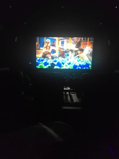 The scene depicts the interior of a darkened cinema auditorium in Tamworth, United Kingdom. A large projection screen centrally displays a vibrant film sequence featuring multiple individuals interacting within a voluminous ball pit composed primarily of blue and green plastic balls. One person with dark, curly hair is partially submerged in the lower left of the screen's content, while another individual with reddish-brown hair is visible in the upper left, also within the ball pit. Several other figures are indistinctly present, engaged in the ball pit environment. The auditorium itself is characterized by low ambient light, with small, recessed light sources visible on the ceiling. Rows of dark cinema seats are discernable in the foreground and midground, appearing as silhouettes against the illumination from the screen. No specific actions or interactions of the audience are clearly visible due to the prevailing darkness.