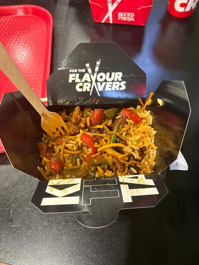 The image shows a close-up, top-down view of a takeaway box filled with food, likely noodles and rice with vegetables and meat. A wooden fork is positioned in the bottom left of the box, about to pick up some food. The box is black with white text that reads "FOR THE FLAVOUR CRAVERS" and also features large white letters spelling out "TA K I". The food appears to be a stir-fry dish with visible pieces of red bell pepper, green beans, and meat. Some of the food has spilled slightly onto the black interior of the box. To the left of the food box, a red tray is visible, and further to the left, parts of another red takeaway container with white text that reads "BO ED FRESH" are visible. The setting appears to be a table, likely in a casual dining environment or at home. The lighting suggests it could be daytime. No people are visible in the image.
