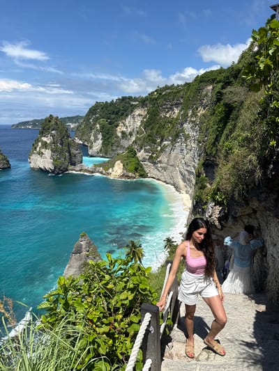 A woman lies on a stone walkway in Pejukutan, Indonesia. She wears a pink tank top, white shorts, and sandals. She leans against a white rope railing. The ocean stretches out below. A cliff face rises at the left. Green vegetation surrounds the scene. The sky is blue with white clouds. Two other people can be seen on the stone walkway in the background.
