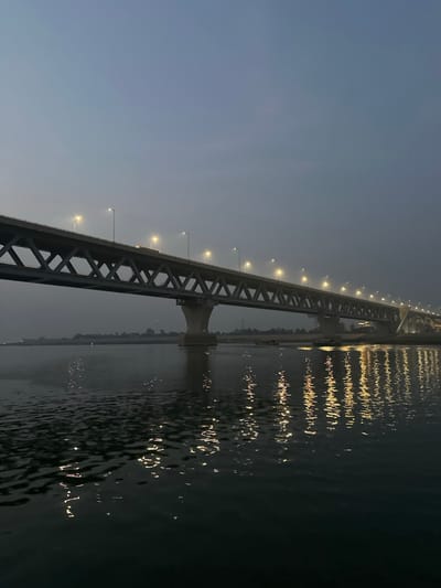 The image displays a segment of the Padma Multipurpose Bridge over a wide river in Naodoba, Bangladesh, observed under low ambient light conditions. The bridge is a multi-span truss structure composed of concrete and steel, supported by visible concrete piers. Numerous evenly spaced, active, warm-toned streetlights illuminate the bridge deck, creating distinct, elongated, wavy reflections on the dark, rippled water surface below. A single vehicle, indistinct in type, is discernible in transit on the illuminated bridge deck. The sky above is a uniform dark grey-blue. Distant, low-lying riverbanks with minimal discernible features are visible to the left and right of the bridge's support structures. Several small, dark objects, consistent with watercraft, are situated near the far riverbank.