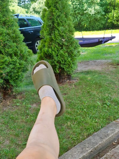 The image shows a person's legs and feet wearing grey socks and olive-green slides, standing on a grassy area. Behind the person, there are evergreen bushes and a black car partially obscured by the foliage. Further in the background, more trees and greenery are visible, along with a glimpse of a purple object that appears to be an umbrella or canopy. A concrete structure, possibly a bench or part of a pathway, is also visible to the right of the frame. The scene is outdoors, likely in a yard or park, with ample sunlight indicating it is daytime. The overall impression is a casual, relaxed setting.