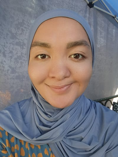 This is a selfie of a woman wearing a blue hijab and a blue and orange patterned top. The woman is smiling and looking at the camera. She has dark brown eyes and dark eyebrows. The background is a gray, textured fabric, possibly a tent or canopy, with some white poles visible at the top. The lighting suggests it is daytime, and the overall scene is casual. The image was taken in Batken City, Kyrgyzstan.