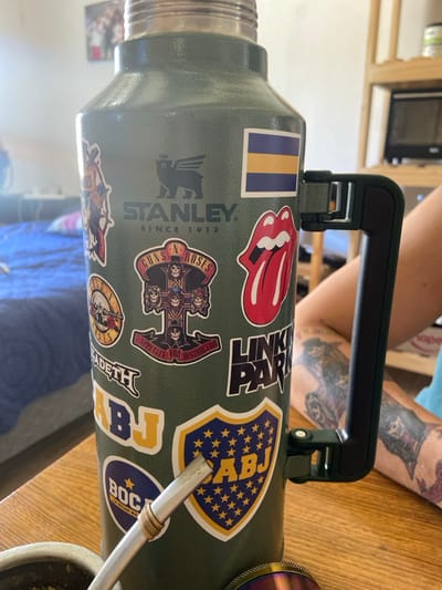 A green Stanley thermos is adorned with stickers. Notable stickers include the Rolling Stones tongue logo, Linkin Park logo, Guns N' Roses Appetite for Destruction album art, and multiple stickers related to the Argentinian football club Boca Juniors. A metal straw, likely for drinking mate, is inserted into the thermos. The background shows a light-colored wooden surface. A tattooed arm is visible in the upper right corner.