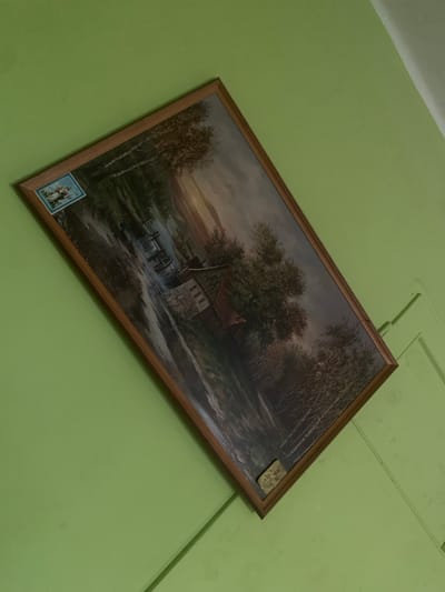 The image shows a framed painting hanging on a light green wall. The painting depicts a pastoral scene with a mill by a river, surrounded by trees. A small, rectangular blue and white religious image is affixed to the upper left corner of the painting's frame. A small, rectangular religious medal is affixed to the lower right corner of the painting's frame. The painting is oriented diagonally in the frame. The room appears to be a domestic interior, possibly a home in Skarydzew, Poland.